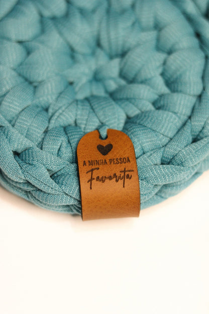 Detalhe da etiqueta de couro gravada com a frase A Minha Pessoa Favorita em base de malha azul.