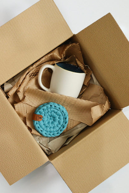 Conjunto de presente com caneca e base em malha azul numa caixa de envio com papel kraft.