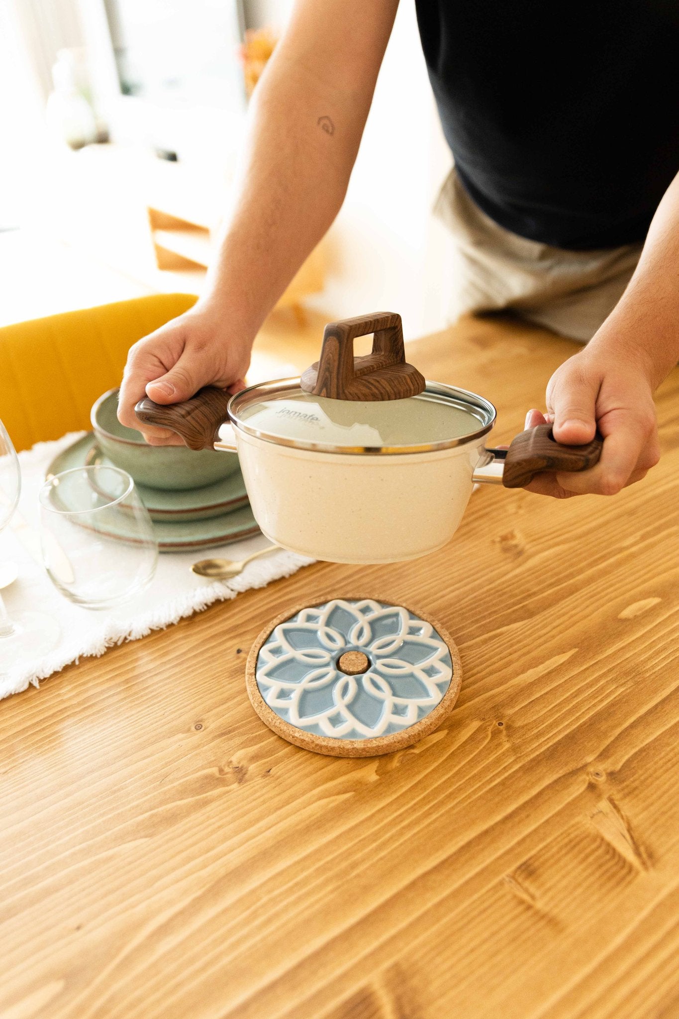 Ambiente de mesa com uma pessoa a colocar uma panela quente sobre a base ALMA azul, demonstrando a funcionalidade e proteção térmica.