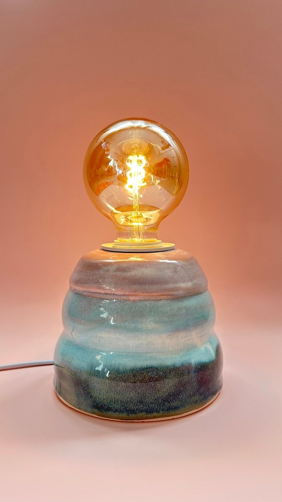 Candeeiro de mesa Aurora em cerâmica grés aceso com luz quente e degradê de tons naturais.