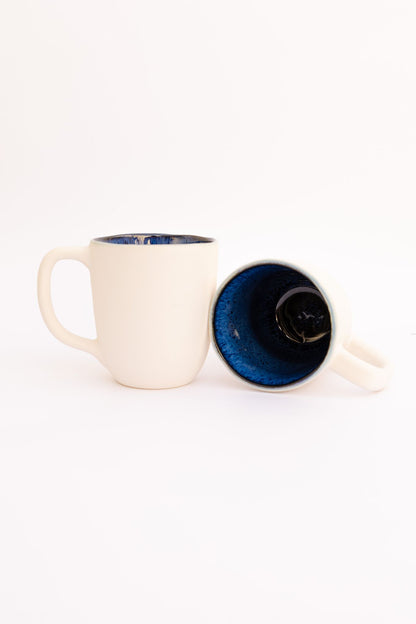 Conjunto de duas canecas CORAL em cerâmica azul e creme, evidenciando o contraste artístico entre o exterior claro e o interior esmaltado reativo.