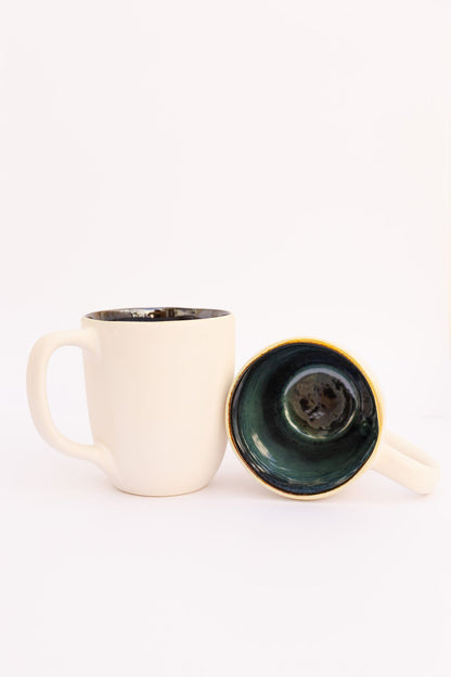 Caneca CORAL verde em cerâmica artesanal, apresentando o contraste entre o exterior creme e o interior verde petróleo vidrado, com uma segunda unidade deitada para detalhe.