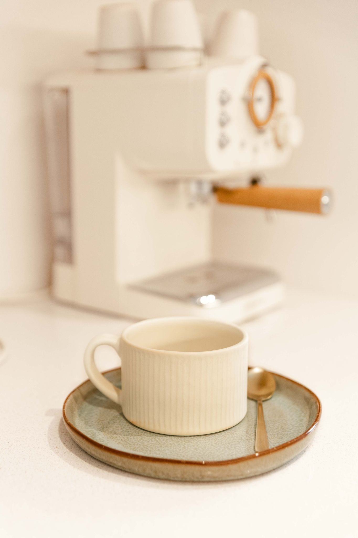 Caneca PLENO bege pousada num pires de cerâmica junto a uma máquina de café expresso, ilustrando um momento de pequeno almoço elegante e moderno.