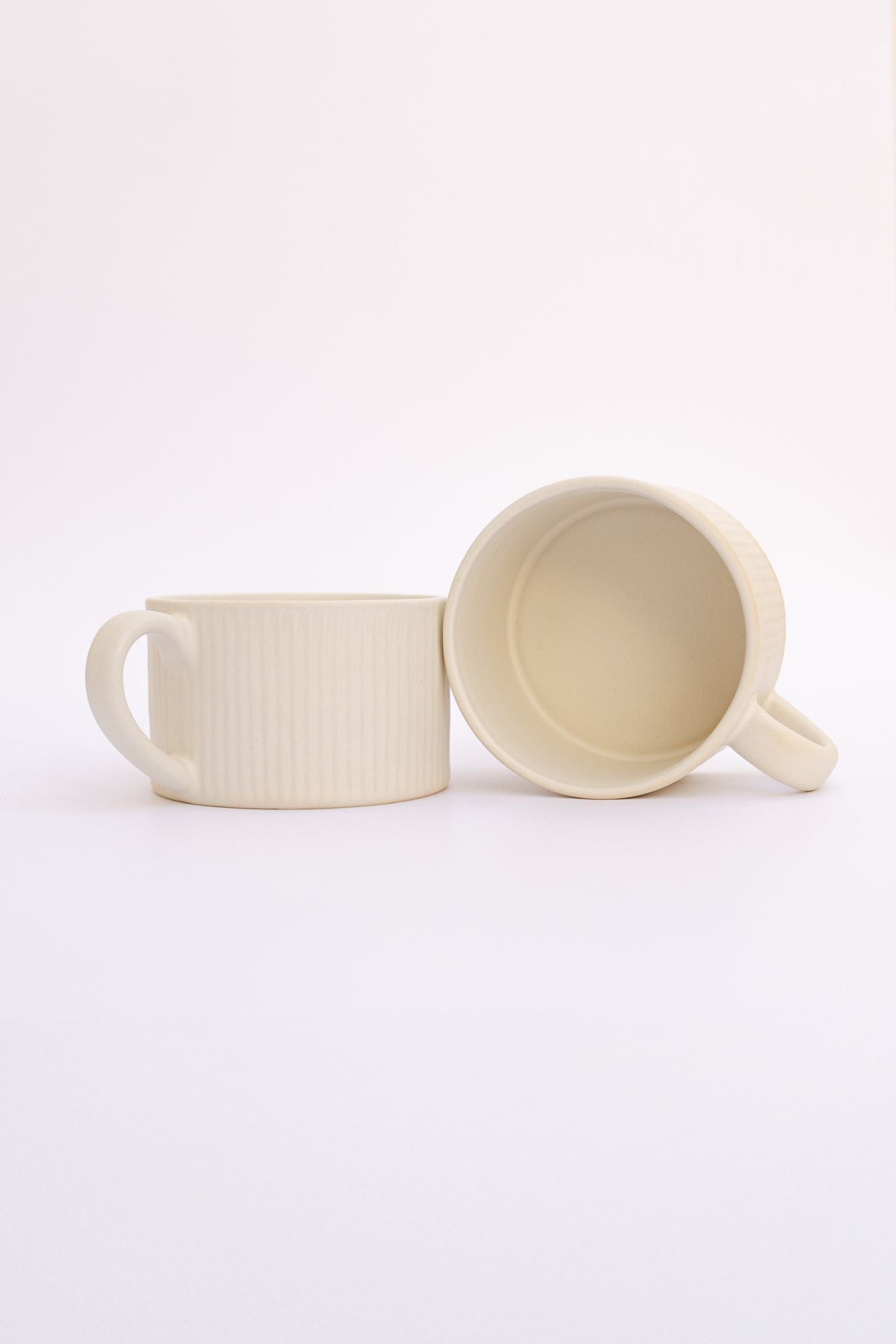 Caneca PLENO em cerâmica bege com textura canelada, apresentada individualmente e com uma segunda unidade deitada para mostrar o interior e o design ergonómico.
