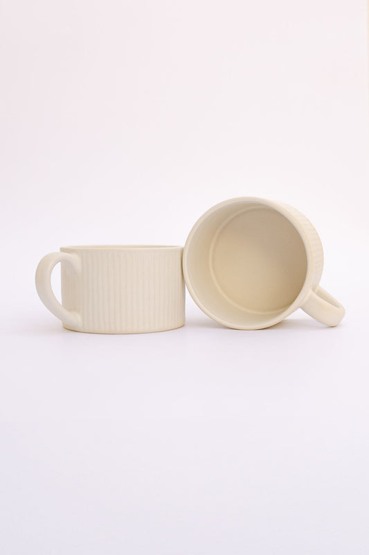 Caneca PLENO em cerâmica bege com textura canelada, apresentada individualmente e com uma segunda unidade deitada para mostrar o interior e o design ergonómico.