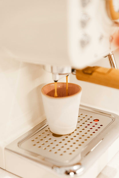 Copo de café CORAL rosa posicionado numa máquina de café expresso de design, capturando o fluxo de café pronto a servir.