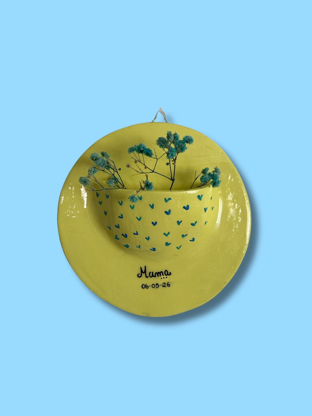 Vaso de parede redondo em cerâmica amarela, personalizado com padrão de pequenos corações azuis e a palavra "Mama" seguida de uma data, pintado à mão.