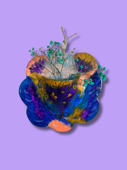 Vaso de parede redondo com borda ondulada em cerâmica, apresentando uma pintura artística abstrata e vibrante em tons de azul, roxo e laranja com flores secas.