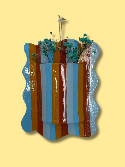 Vaso de parede retangular personalizado em cerâmica fria, com borda ondulada, padrão de riscas coloridas em azul, amarelo e castanho e flores decorativas.